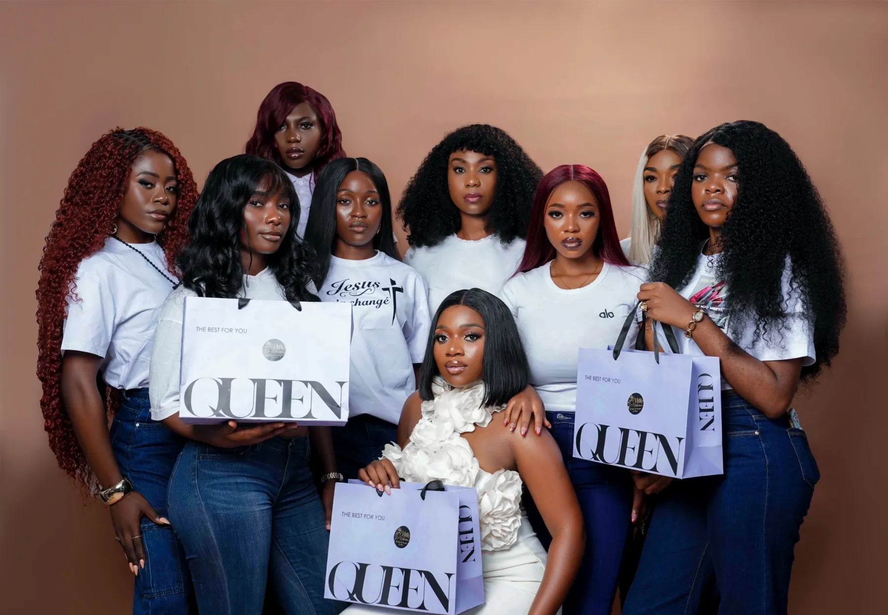 Nine women from the VMK family wearing different VMK hair — copper kinky curls, black body wave, burgundy, platinum blonde and a sleek bob — holding lilac QUEEN bags